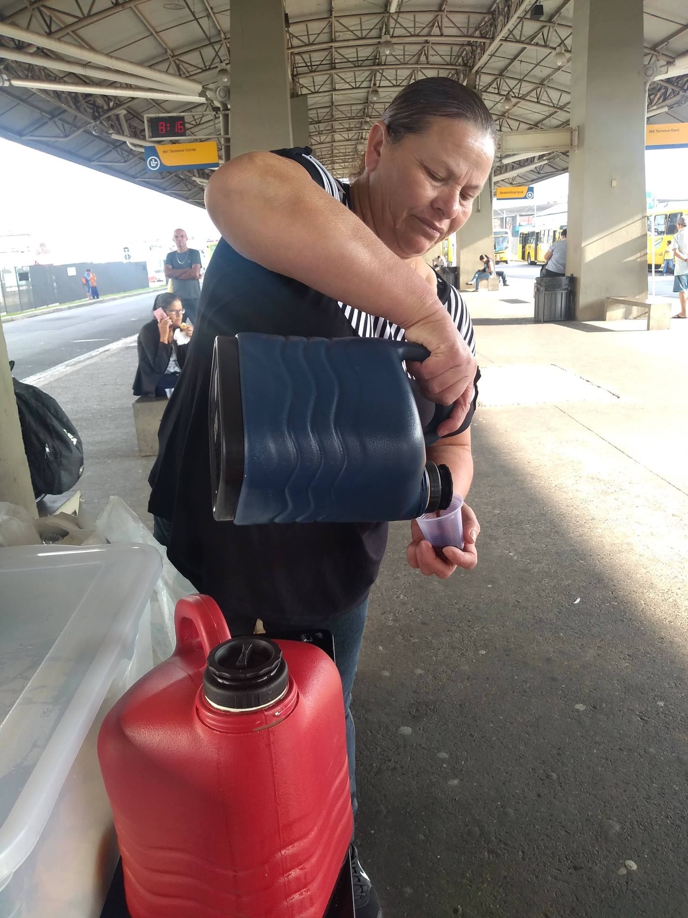 A mãe do Cláudio trabalhando no terminal de ônibus, servindo café de uma garrafa térmica; o relógio da plataforma marca 8:16